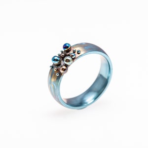 Può includere: Un anello in titanio blu con un gruppo di piccole perle di metallo iridescente sulla parte superiore.