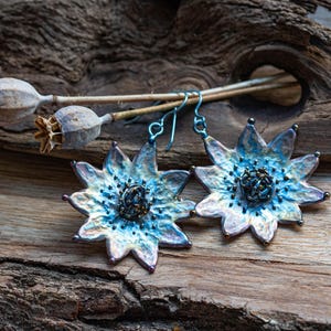 Puede incluir: Un par de pendientes en forma de flor azul e iridiscente con un centro de cuentas. Los pendientes cuelgan de una superficie de madera.