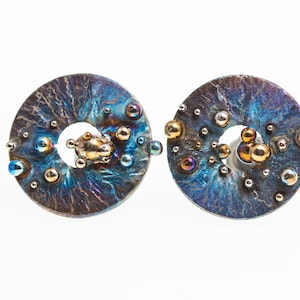 May include: A pair of round, silver earrings with a textured surface and a ring in the center. The earrings are decorated with small, iridescent blue and gold beads.