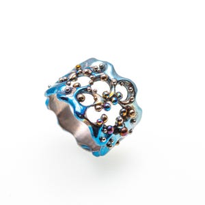 May include: A blue and silver ring with a delicate, intricate design. The ring is made of metal and has a textured surface. The ring is decorated with small, round beads.