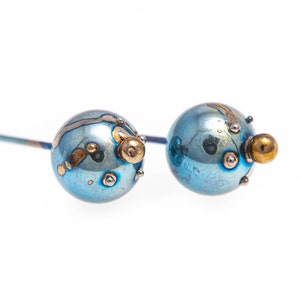 May include: A pair of blue titanium earrings with gold accents. The earrings are round and have a textured surface.