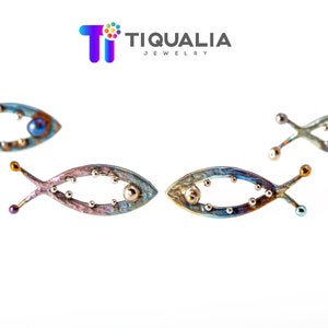 May include: Four fish-shaped jewelry pieces with iridescent blue, purple, and gold tones are displayed against a white background. The jewelry has small, round accents and the text "TIQUALIA JEWELRY" is visible.