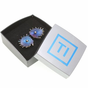 Op de afbeelding: Een paar blauwe en paarse titanium oorbellen met een stervormig ontwerp, in een zilveren doos met de letter "TI" in blauw.
