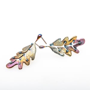 May include: A pair of earrings shaped like oak leaves with a metallic finish. The leaves are a mix of gold, copper, and silver tones with small blue beads scattered throughout.