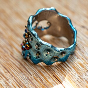 May include: A blue metal ring with a textured surface and small silver and gold beads. The ring is on a wooden surface.