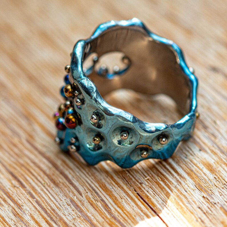 May include: A blue metal ring with a textured surface and small silver and gold beads. The ring is on a wooden surface.