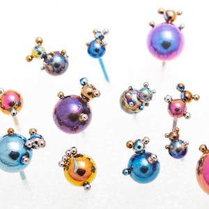 May include: A collection of iridescent, metallic, sphere-shaped ornaments with small, decorative details. The ornaments are arranged on a white surface and are attached to thin, silver wires.