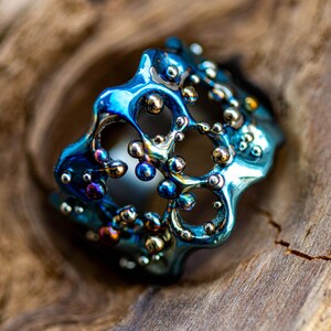 May include: A blue and gold metal ring with a unique, organic design. The ring is made of a textured metal with a bubble-like pattern.