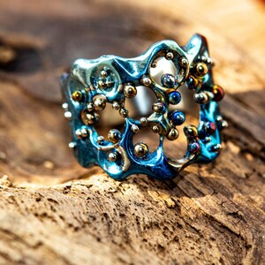 May include: A blue and gold ring with a unique, organic design. The ring is made of metal and has a textured surface. The ring is set with small, round beads.