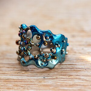 May include: A blue metal ring with a unique, organic design featuring small, round, silver and gold accents. The ring has a textured, uneven surface.