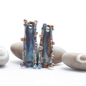 K&ouml;nnte beinhalten: Ein Paar regenbogenfarbene blaue und goldene Ohrringe mit einer strukturierten, geh&auml;mmerten Metalloberfl&auml;che. Die Ohrringe sind mit kleinen, runden Perlen verziert.