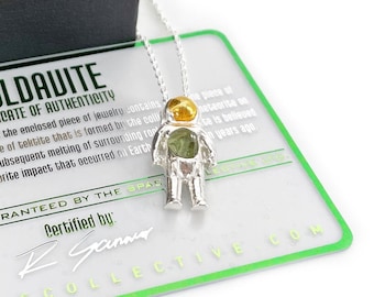 May include: A silver astronaut pendant necklace with a green and yellow moldavite stone. The necklace comes with a certificate of authenticity that states "Moldavite is a type of tektite that is formed by the collision of a meteorite with the Earth's surface resulting in the subsequent melting of surrounding rock." The certificate is signed by "K. Gamma" and includes the logo "THESPACECOLLECTIVE.COM".