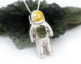 May include: A silver astronaut pendant with a yellow gemstone in the helmet. The pendant is hanging from a silver chain and is resting on a green rock.