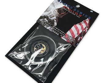 May include: A small, round, black disc with a silver Apollo 17 logo and the text "Flown from NASA's Famous Missions" in white. The disc is packaged in a clear plastic bag with a black card backing that reads "The Space Series Apollo 17 Flown Kapton Foil".