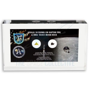 Può includere: Una teca bianca con un'immagine in bianco e nero della missione Apollo 10. Il testo "APOLLO 10 FLOWN CM KAPTON FOIL & NWA 10203 MOON ROCK" è stampato sull'immagine. Ci sono due piccoli contenitori rotondi e trasparenti con un pezzo di roccia lunare e un pezzo di foglio di Kapton.