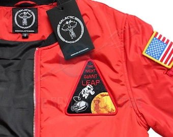 May include: Red bomber jacket with a black zipper and a black and white patch that says "The Next Giant Leap" with an astronaut and a planet. The jacket also has a small American flag patch on the right sleeve.