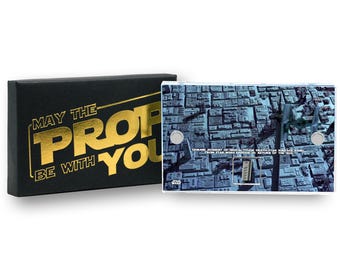 May include: A black box with gold text that reads "MAY THE PROP BE WITH YOU" next to a rectangular item with a Death Star surface design. The item has two silver circular fasteners and text that reads "GENUINE ELEMENT OF HIGH ALTITUDE DEATH STAR SURFACE PANEL FROM STAR WARS EPISODE VI: RETURN OF THE JEDI."