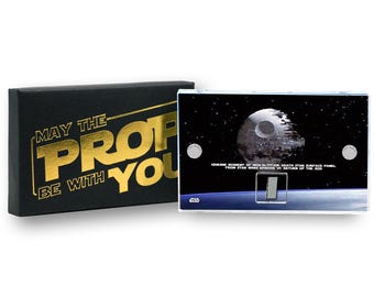 May include: A Star Wars collectible featuring a Death Star surface panel. The panel is displayed against a space backdrop. The black box next to it has the text "MAY THE PROF BE WITH YOU" in gold.