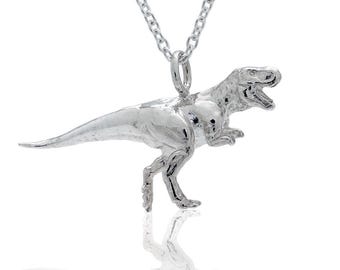 May include: A silver-colored T-Rex pendant necklace. The dinosaur is in a walking pose with its mouth open. The necklace chain is a silver-colored curb chain.