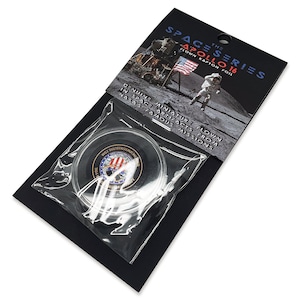 Può includere: Una piccola moneta rotonda d'argento con un design rosso, bianco e blu è esposta in una custodia di plastica trasparente. La moneta è etichettata "A COLLECTION E.COM" e "FLOWN KAPTON FOIL". Lo sfondo è nero con un'etichetta bianca che recita "THE SPACE SERIES APOLLO 16".