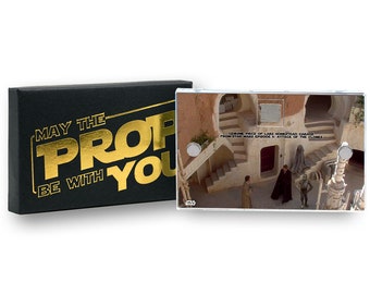May include: A clear plastic frame with a photo of a scene from Star Wars Episode II: Attack of the Clones. The photo shows a building exterior with a staircase and three figures standing in the courtyard. The text "GEMINE PIECE OF LARS HOMESTEAD GARAGE FROM STAR WARS EPISODE II: ATTACK OF THE CLONES" is printed on the frame.