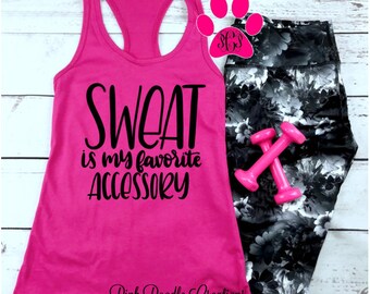 May include: Pink tank top with the text "SWEAT is my favorite ACCESSORY" in black. The tank top is laying on a white wooden surface with black and white floral leggings, pink dumbbells, and a pink paw print with the letters "SCS" in the center.