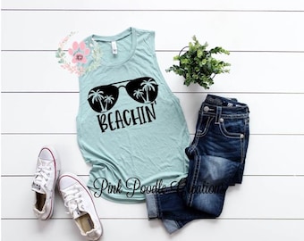 May include: A light blue sleeveless tank top with a black graphic of sunglasses and palm trees. The text "Beachin'" is printed below the sunglasses.