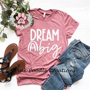 May include: A pink t-shirt with the text "DREAM big" in white lettering. The shirt has a rainbow graphic with a heart in the center. The shirt is laid out on a white surface with a pair of sandals and a pair of jeans.