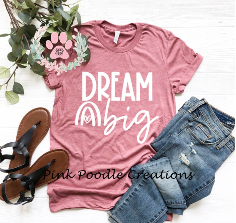 May include: A pink t-shirt with the text "DREAM big" in white lettering. The shirt has a rainbow graphic with a heart in the center. The shirt is laid out on a white surface with a pair of sandals and a pair of jeans.