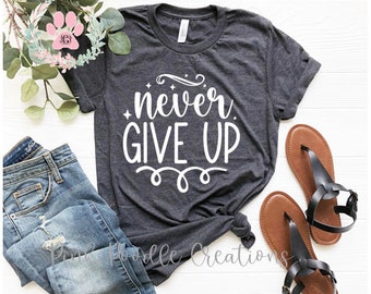 May include: A dark grey t-shirt with the text "never give up" in white lettering. The shirt is styled with a pair of blue jeans and brown sandals.