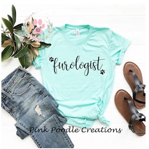 May include: A light turquoise t-shirt with the word "furoglogist" in black script, accompanied by paw print graphics. The shirt is styled with blue jeans, sandals, and floral accents, creating a casual, pet-themed aesthetic. The text "Pink Poodle Creations" is at the bottom.