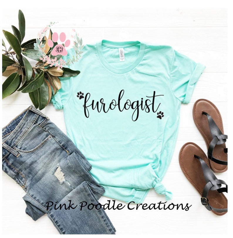 May include: A light turquoise t-shirt with the word "furoglogist" in black script, accompanied by paw print graphics. The shirt is styled with blue jeans, sandals, and floral accents, creating a casual, pet-themed aesthetic. The text "Pink Poodle Creations" is at the bottom.