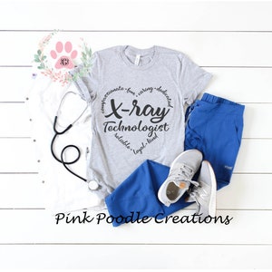 May include: A gray t-shirt with a black circle design that says "Compassionate - Fun - Caring - Dedicated - X-ray Technologist - Reliable - Loyal - Kind". The shirt is laying on a white surface with a stethoscope, blue scrubs, and gray sneakers.