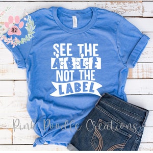 May include: A blue t-shirt with white text that reads "See the Puzzle Not the Label". The text is in a puzzle piece font. The shirt is laying on a white wooden surface with a pair of blue jeans.