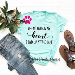 Puede incluir: Una camiseta azul turquesa con el texto "WHEN I FOLLOW MY heart I END UP AT THE LAKE" en negro. Una huella de pata rosa con un monograma est&aacute; en el lado izquierdo de la camiseta. La camiseta est&aacute; colocada sobre una superficie blanca con un par de sandalias marrones y unos vaqueros azules.