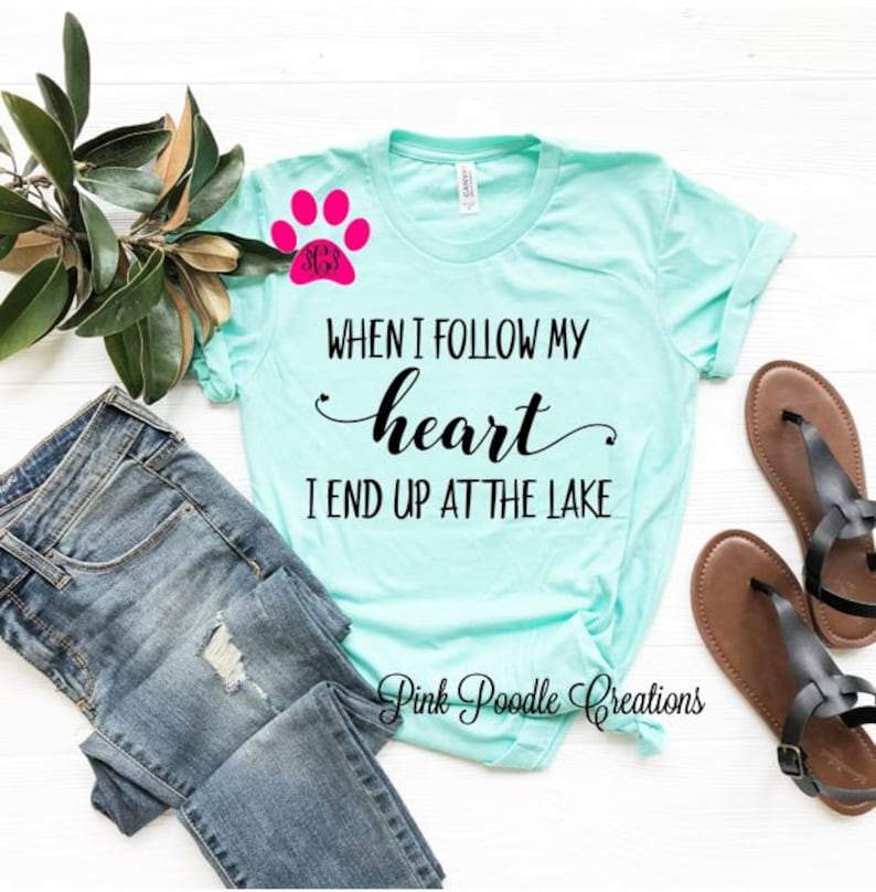 Puede incluir: Una camiseta azul turquesa con el texto "WHEN I FOLLOW MY heart I END UP AT THE LAKE" en negro. Una huella de pata rosa con un monograma est&aacute; en el lado izquierdo de la camiseta. La camiseta est&aacute; colocada sobre una superficie blanca con un par de sandalias marrones y unos vaqueros azules.