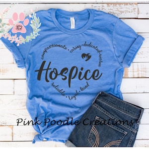 May include: A blue t-shirt with a white print that says "Hospice" in a circle with the words "compassionate, caring, dedicated, warm, reliable, loyal, kind" around it. The shirt is on a white wooden surface with a pair of blue jeans.