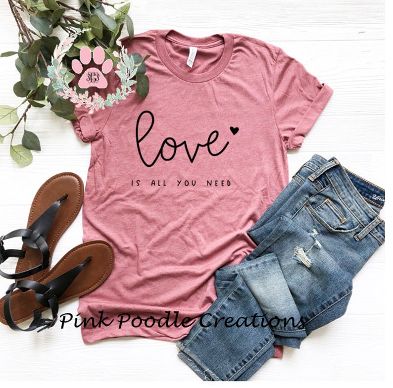 May include: A dusty rose t-shirt with the words "love is all you need" in black script. The shirt is paired with blue jeans and brown sandals. A floral wreath and the text "Pink Poodle Creations" are also visible.