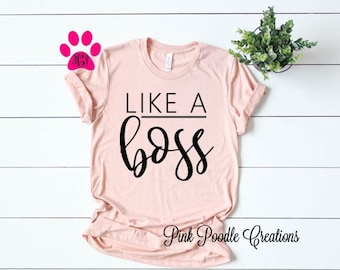 May include: A pink t-shirt with the text "Like a Boss" in black lettering. The shirt is on a white wooden background.