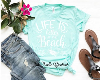 May include: A turquoise t-shirt with the text "Life is better at the beach" in white. The shirt has a white graphic of a turtle and starfish. The text "Pink Poodle Creations" is at the bottom of the shirt.