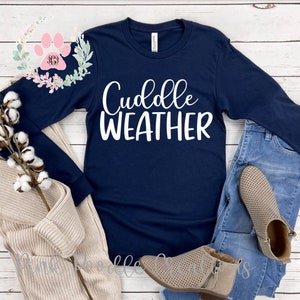 May include: Navy blue long-sleeve shirt with the text "Cuddle Weather" in white. The shirt is displayed on a white wooden surface with a pair of jeans and brown boots.