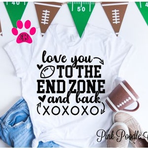 May include: White t-shirt with black text that reads "love you to the end zone and back xoxo". A football graphic is included in the text. The shirt is laying on a blue jean fabric and a brown and white football-shaped mug.