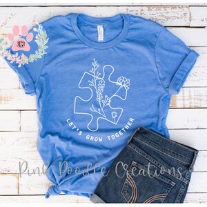 May include: A blue t-shirt with a white outline of a puzzle piece with flowers growing inside. The text "Let's Grow Together" is written around the puzzle piece.