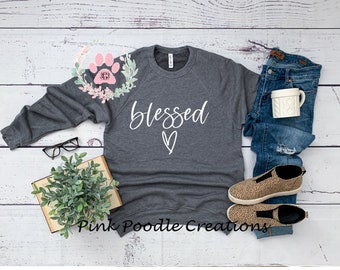 May include: A gray sweatshirt with the word "blessed" in white cursive lettering and a white heart underneath. The sweatshirt is laid flat on a white wooden surface with a pair of glasses, a book, a potted plant, a pair of jeans, a coffee mug, and a pair of leopard print shoes.