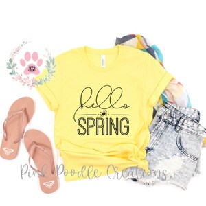 May include: Yellow t-shirt with black text that says "hello spring" with a sun graphic. The shirt is folded and laid on a white surface with a pair of brown flip flops, a pair of denim shorts, and a colorful scarf.