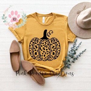 May include: A mustard yellow t-shirt with a black leopard print pumpkin design. The shirt is laying flat on a white surface with a pair of brown shoes and a brown hat.