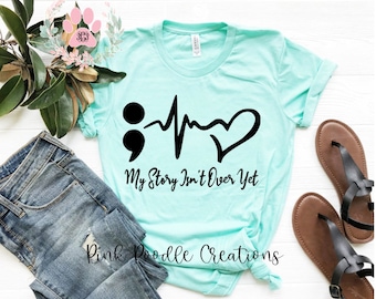 Peut inclure: Un t-shirt turquoise avec un point-virgule, un rythme cardiaque et un cœur noirs. Le texte "My Story Isn't Over Yet" est imprimé sous le graphique. Le t-shirt est posé à plat sur une surface en bois blanc.