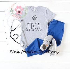 May include: A gray t-shirt with the text "MEDICAL assistant" and a small potted plant graphic. The shirt is laid out on a white surface with a stethoscope, blue scrubs, and gray sneakers.