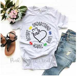 May include: A gray t-shirt with a circle design featuring a heart with a puzzle piece inside. The words "Accept, Teach, Love, Understand" are written around the heart in a circle. The puzzle pieces are in the colors red, yellow, green, and blue.