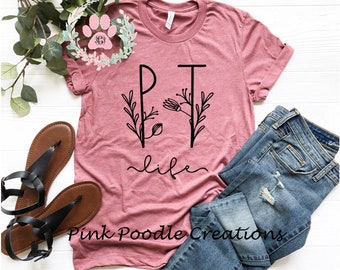 May include: A pink t-shirt with the text "PT life" in black lettering. The letters are surrounded by floral designs. The shirt is laid out on a white surface with a pair of brown sandals and blue jeans.